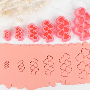 May include: A set of six pink heart-shaped cookie cutters, arranged in order of size, with the largest cutter at the top and the smallest cutter at the bottom. The cutters are on a white surface, with a pink fondant sheet below them. The fondant sheet has been cut with the cookie cutters to create a row of heart-shaped impressions.