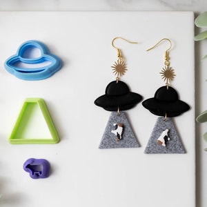 Può includere: Un paio di orecchini pendenti con UFO neri con dettagli a stella dorati e ciondoli triangolari grigi con figure di mucche bianche. Gli orecchini sono esposti su una superficie bianca con tagliabiscotti blu, verdi e viola.