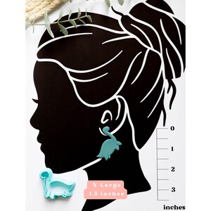 May include: A teal blue dinosaur-shaped earring with a measurement guide showing the earring is 1.3 inches in size. The text "X-Large 1.3 inches" is printed on a pink background.