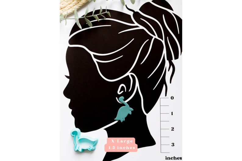 May include: A teal blue dinosaur-shaped earring with a measurement guide showing the earring is 1.3 inches in size. The text "X-Large 1.3 inches" is printed on a pink background.