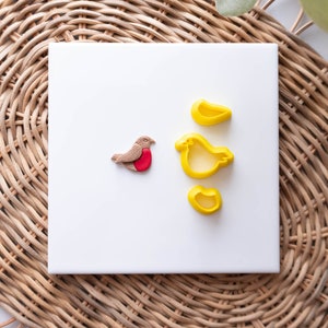 May include: A white ceramic tile with a red and brown robin cutout and three yellow bird-shaped cookie cutters.