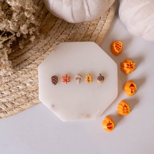 May include: A white marble tile with six small autumn-themed clay charms arranged in a row. The charms include an acorn, a maple leaf, a mushroom, an oak leaf, and two acorns. The tile is surrounded by a woven straw mat and several orange clay charms.