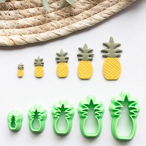 Könnte beinhalten: Ein Set mit sechs grünen Ananas-Ausstechformen und sechs gelben und grünen Ananas-Fondant-Dekorationen. Die Ausstechformen sind in verschiedenen Größen und die Dekorationen sind in einer Reihe von klein nach groß angeordnet.