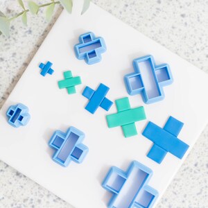 May include: A set of nine plastic cookie cutters in the shapes of plus signs. The cutters are in various sizes and colours, including blue and green.