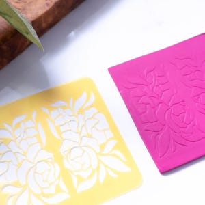 May include: Two floral stencils, one yellow and one pink. The yellow stencil is a cut-out design, while the pink stencil is raised and embossed.