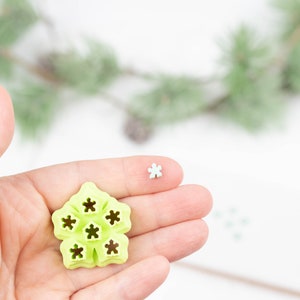 Può includere: Un tagliapasta a forma di fiocco di neve in plastica verde a sei punte. Il tagliapasta è tenuto in una mano, ed è visibile anche un fiocco di neve bianco più piccolo.