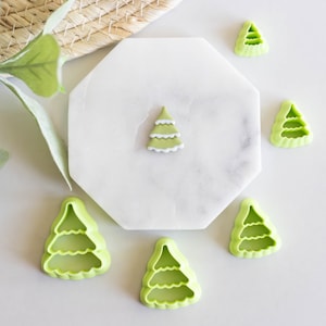Könnte beinhalten: Grüne Plastik-Ausstecher in Form von Weihnachtsbäumen auf einer weißen Oberfläche. Einer der Ausstecher ist mit grünem und weißem Zuckerguss gefüllt.