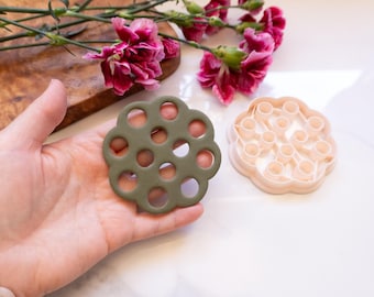 Puede incluir: Un cortador de galletas verde y circular con múltiples agujeros se sostiene en una mano. Un segundo cortador de galletas rosa claro está al lado. Flores rosas y una tabla de cortar de madera están en el fondo. Los cortadores de galletas son para hornear.