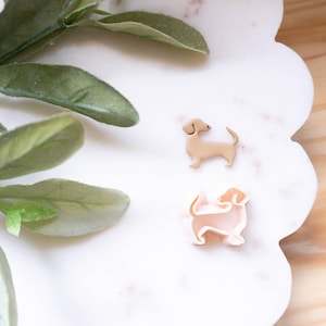 Puede incluir: Dos pendientes con forma de perro salchicha en tonos marrón claro y rosa descansan sobre una superficie blanca festoneada. Los pendientes son pequeños y detallados, con características visibles. Hojas y tallos verdes en la esquina superior izquierda.