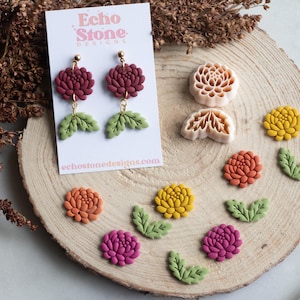 Op de afbeelding: Een paar kastanjebruine bloemoorbellen met groene blaadjes op een witte Echo Stone Designs-kaart, vergezeld door twee beige plastic bloem- en bladvormpjes en diverse losse kleivormen in de kleuren geel, oranje en roze.
