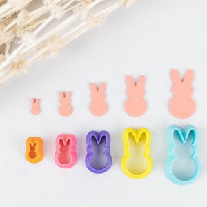 May include: A set of nine bunny-shaped cookie cutters in various sizes and colors, including orange, pink, purple, blue, and yellow. The cutters are arranged in a row on a white surface.