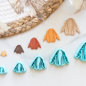 May include: A set of eight cookie cutters in various sizes, shaped like sweaters. The cutters are made of blue plastic and have a detailed design.