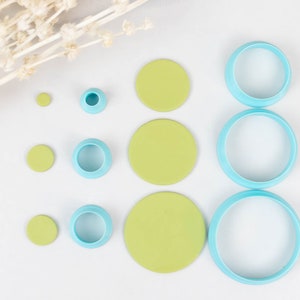 May include: A set of eight round cookie cutters in various sizes. The cutters are light blue and have a smooth, shiny finish. The cutters are arranged in a row on a white surface, with the largest cutter at the right and the smallest cutter at the left. There are also five round green circles of various sizes on the surface.