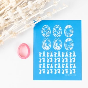 Pode incluir: Uma folha de estêncil azul com desenhos brancos de ovos de Páscoa e coelhos. A folha de estêncil está sobre uma superfície branca com um anel rosa ao lado.