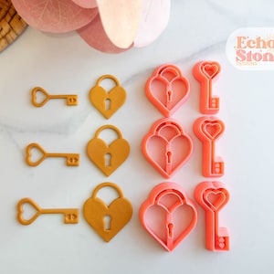 Peut inclure: Neuf emporte-pièces en forme de cœur dorés et roses, dont trois emporte-pièces en forme de clé et six emporte-pièces en forme de cadenas. Les emporte-pièces sont disposés en trois rangées sur une surface blanche. Le texte "Echo Stone Designs" est visible dans le coin supérieur droit de l'image.
