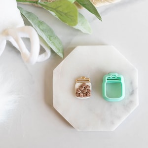 May include: A jar-shaped polymer clay charm filled with small brown balls, next to a mint green jar-shaped cookie cutter. The charm has a gold-coloured lid. The items are on a white, octagonal surface.