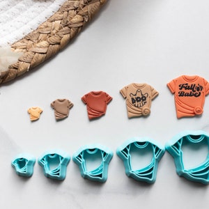 May include: Set of six cookie cutters in the shape of t-shirts. The cutters are blue and come in various sizes. There are also three small t-shirts made of fondant in brown, orange, and tan.