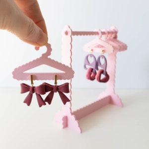 Op de afbeelding: Een roze miniatuur kledingrek met drie hangers met paren van bordeauxrode strik oorbellen.