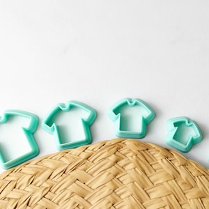 May include: Five teal blue plastic cookie cutters shaped like t-shirts, arranged in a row from largest to smallest on a woven straw surface.