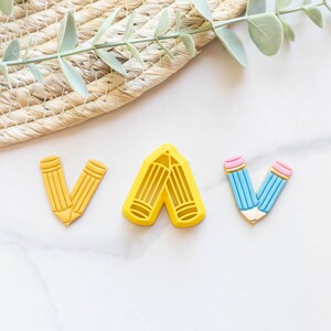 May include: Three pencil-shaped cookie cutters, one yellow, one blue and pink, and one yellow. The cutters are arranged in a row on a white marble surface.