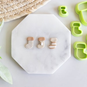 May include: Three small, hand-crafted Christmas stocking ornaments in white, brown, and tan with different patterns. The ornaments are on a white marble surface.