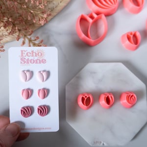 Op de afbeelding: Een set van zes roze tulpenvormige oorbellen op een witte kaart met de tekst "Echo Stone Designs" en een websiteadres. De oorbellen zijn gerangschikt in drie paar, waarbij elk paar een iets andere roze tint heeft.