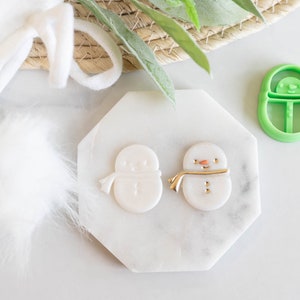May include: Two white fondant snowman ornaments with gold details, one with a red nose and the other without, on a white marble surface. A green plastic snowman cookie cutter is in the background.