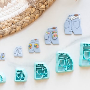 Puede incluir: Un conjunto de nueve cortadores de galletas de plástico azul y turquesa con forma de bolsillos traseros de jeans. Los cortadores están dispuestos en orden de tamaño, con el cortador más grande en la parte superior y el más pequeño en la parte inferior.