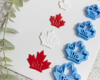 Può includere: Un set di sei tagliapasta a forma di foglia d'acero canadese. I tagliapasta sono in rosso, bianco e blu. I tagliapasta hanno la forma di una foglia d'acero e hanno la scritta "eh?" o "eh!" su di essi.