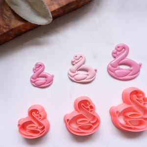 Puede incluir: Cortadores de galletas rosas con forma de flamenco en varios tamaños. Los cortadores son en tonos de rosa, de claro a oscuro, y están dispuestos sobre una superficie blanca. Los cortadores son de plástico y están diseñados para hornear.