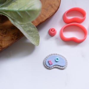 Puede incluir: Una colección de cortadores de arcilla polimérica de color coral y una creación de arcilla en miniatura. Los cortadores incluyen dos formas de riñón, un pequeño cilindro y una pieza en forma de rosa. La creación en miniatura es una piscina azul y rosa.