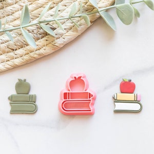 Puede incluir: Tres cortadores de galletas con forma de pila de libros con una manzana en la parte superior. Los cortadores son de color rosa, verde y amarillo.
