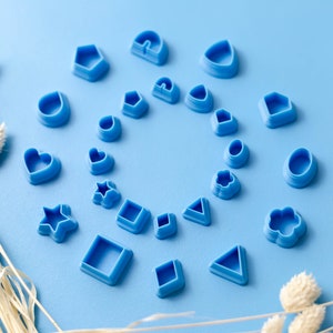 May include: A collection of blue plastic cookie cutters in various shapes, including hearts, stars, flowers, and geometric shapes, arranged in a circle on a light blue background.