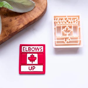 Könnte beinhalten: Roter und weißer Ausstecher mit den Worten "Elbows Up" und einem kanadischen Flaggen-Design.