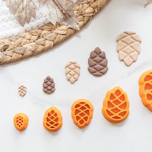 May include: A set of nine different sized pine cone shaped polymer clay charms in shades of brown, orange, and beige.