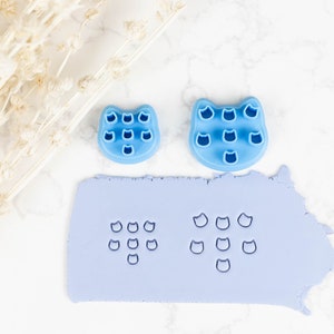 May include: Two blue plastic cat-shaped cookie cutters with multiple cat face impressions. The larger cutter is on the right and the smaller cutter is on the left. A sheet of light blue fondant has been pressed with the cutters, creating multiple cat face impressions.