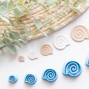 Può includere: Un set di 9 tagliapasta a forma di conchiglia di diverse dimensioni. I tagliapasta sono in plastica blu e bianca. I tagliapasta sono disposti in fila su una superficie bianca.