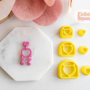 Può includere: Un set di stampi per orecchini in argilla polimerica a forma di cuore con un ritaglio. Gli stampi sono gialli e sono mostrati con un orecchino in argilla polimerica rosa che è stato realizzato usando gli stampi. Il testo "Echo Stone Designs" è visibile nell'angolo in alto a destra dell'immagine.