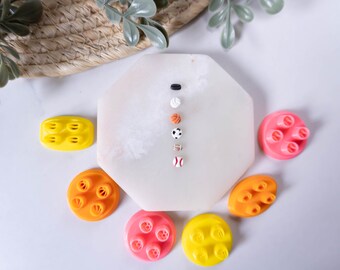 May include: A set of six miniature sports ball charms, including a baseball, basketball, football, soccer ball, volleyball, and a black and white striped golf ball. The charms are arranged vertically on a white octagonal plate. The plate is surrounded by four colorful plastic molds.