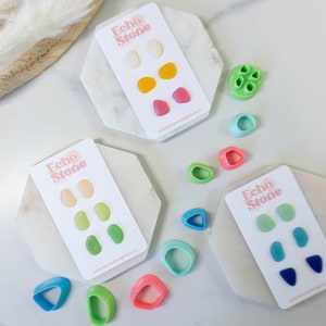 Könnte beinhalten: Drei Sätze farbenfroher Polymer Clay Ohrringe auf weißen Karten mit dem Text "Echo Stone" und einer Sonnengrafik. Die Ohrringe sind in Grün-, Blau- und Rosatönen.