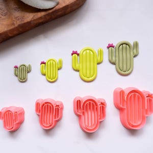 Puede incluir: Una colección de cortadores de galletas con forma de cactus en varios tamaños y colores. Los cortadores son en tonos verdes, amarillos y rosas, con pequeños acentos de flores rosas. Los cortadores están dispuestos sobre una superficie blanca.