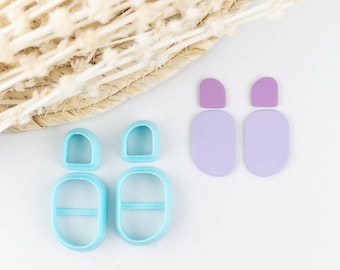 Könnte beinhalten: Zwei hellblaue Ohrring-Ausstechformen aus Kunststoff mit abgerundetem Bogen oben und einer größeren ovalen Basis, dazu ein Paar passende violette Ohrringe, bestehend aus einem kleinen halbkreisförmigen Oberteil und einem größeren ovalen Unterteil.