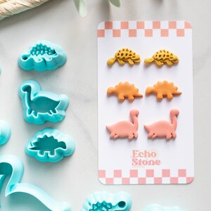 May include: A set of six dinosaur-shaped cookie cutters in a light blue color. The cutters are arranged in a row on a white surface. Next to the cutters is a card with six dinosaur-shaped cookies in yellow, orange, and pink colors. The card has the text "Echo Stone" printed on it.