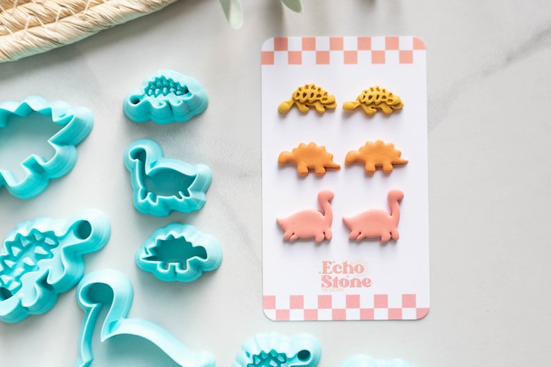 May include: A set of six dinosaur-shaped cookie cutters in a light blue color. The cutters are arranged in a row on a white surface. Next to the cutters is a card with six dinosaur-shaped cookies in yellow, orange, and pink colors. The card has the text "Echo Stone" printed on it.