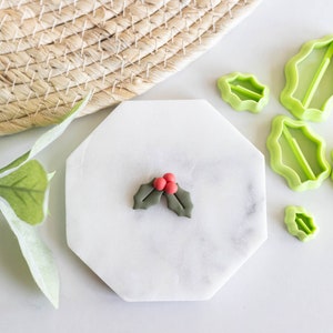 Puede incluir: Cortadores de galletas de hoja de acebo de plástico verde con una baya de acebo roja sobre una superficie de mármol blanco.