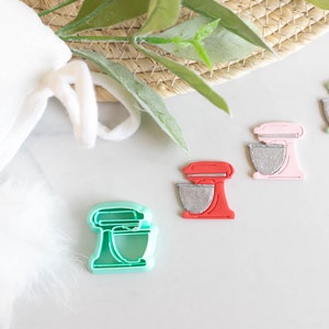 May include: Four different colored cookie cutters shaped like a stand mixer. The cookie cutters are green, pink, red, and teal.