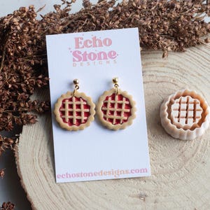 May include: A pair of dangle earrings shaped like miniature cherry pies with a tan crust and red filling, attached to a white display card labelled Echo Stone Designs. Beside the card is a matching circular pie-shaped plastic cutter.