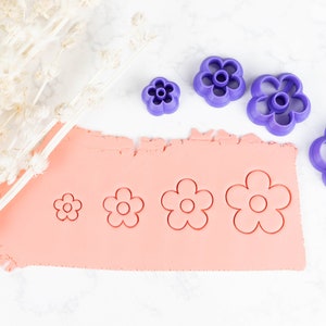 May include: Four purple plastic flower-shaped cookie cutters of different sizes on a white surface. The largest cutter is on the right side of the image. The three smaller cutters are on the left side of the image. A pink sheet of clay is in the background with the cookie cutters pressed into it.
