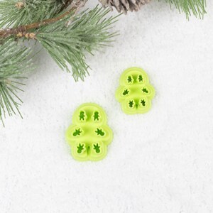 May include: Two green plastic cookie cutters shaped like Christmas trees on a white background. The cookie cutters are made of a flexible plastic and have a snowflake design.
