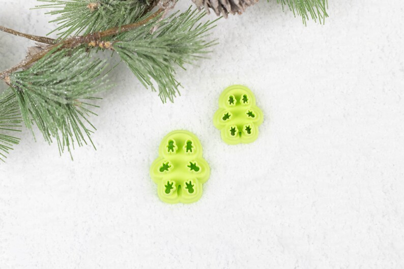 May include: Two green plastic cookie cutters shaped like Christmas trees on a white background. The cookie cutters are made of a flexible plastic and have a snowflake design.